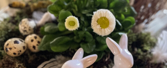 Zwei Osterhasen aus Porzellan stehen vor einem österlichen Gesteck.