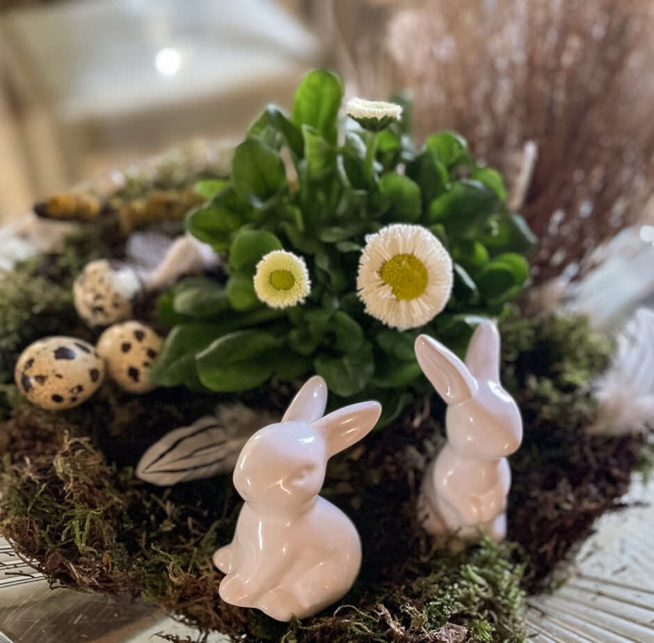 Zwei Osterhasen aus Porzellan stehen vor einem österlichen Gesteck.