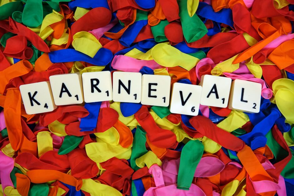 Scrabble-Buchstaben formen das Wort "Karneval" und im Hintergund liegen bunte Luftballons, die noch nicht aufgeblasen sind.
