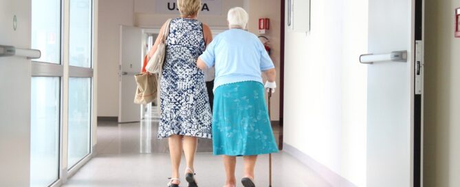 Zwei Frauen laufen untergehakt durch einen Krankenhausflur.