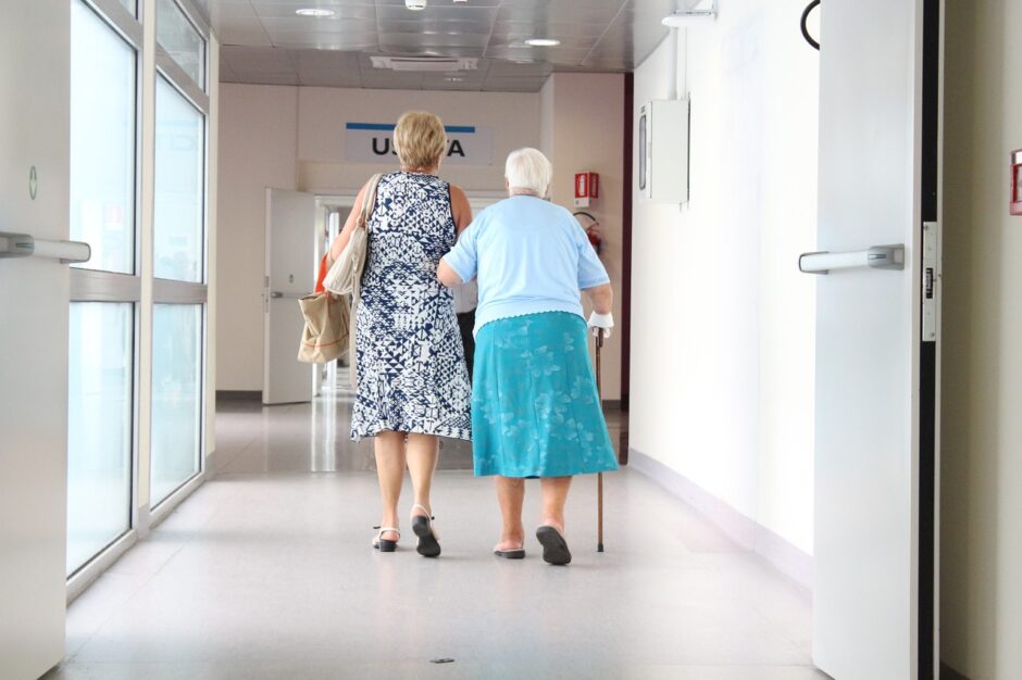 Zwei Frauen laufen untergehakt durch einen Krankenhausflur.