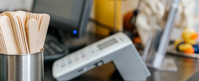 Schreibtisch eines Arztes mit Holzstäbchen um den rachenraum zu überprüfen, einem Skelett der Wirbelsäule im Hintergrund, ein Versicherungskarten-Lesegerät und einem PC im Hintergrund.