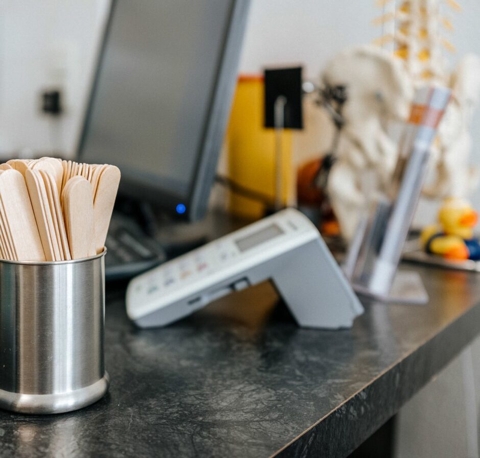Schreibtisch eines Arztes mit Holzstäbchen um den rachenraum zu überprüfen, einem Skelett der Wirbelsäule im Hintergrund, ein Versicherungskarten-Lesegerät und einem PC im Hintergrund.