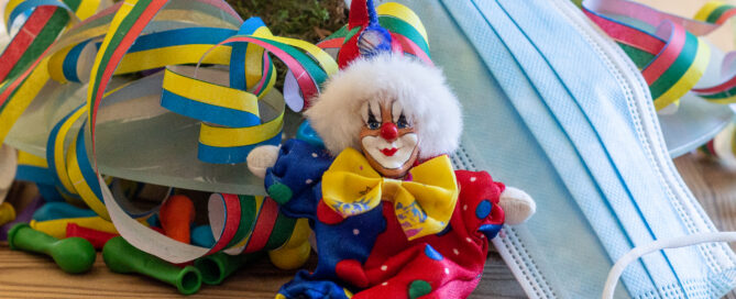 Ein Mundschutz, ein Porzellan-Clown, Luftschlangen sowie Luftballons liegen auf einem Tisch.