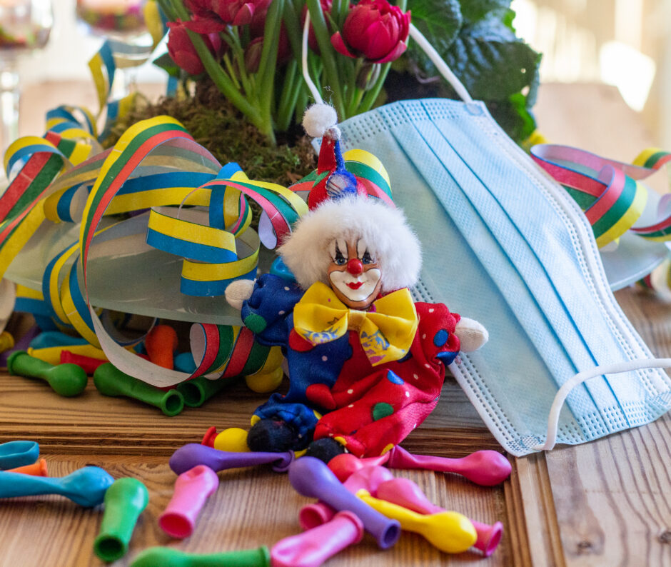 Ein Mundschutz, ein Porzellan-Clown, Luftschlangen sowie Luftballons liegen auf einem Tisch.