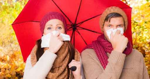 Ein mann und eine Frau stehen Naseputzend unter einem roten Regenschirm vor Herbstbäumen.