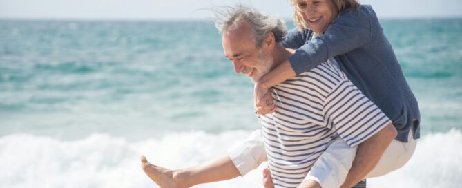 Ein Mann trägt eine Frau Huckepack auf dem Rücken am Strand im Sand entlang.