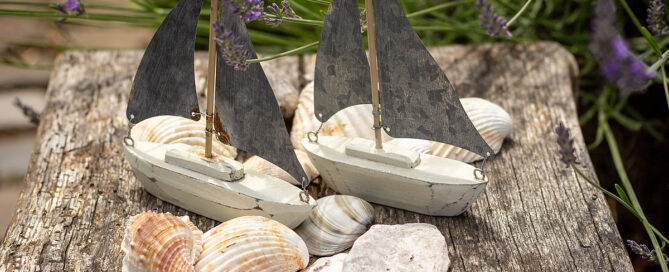 Ein sommerliches Deko-Arrangement aus zwei Segelbooten auf einem Holztisch und Muscheln davor.