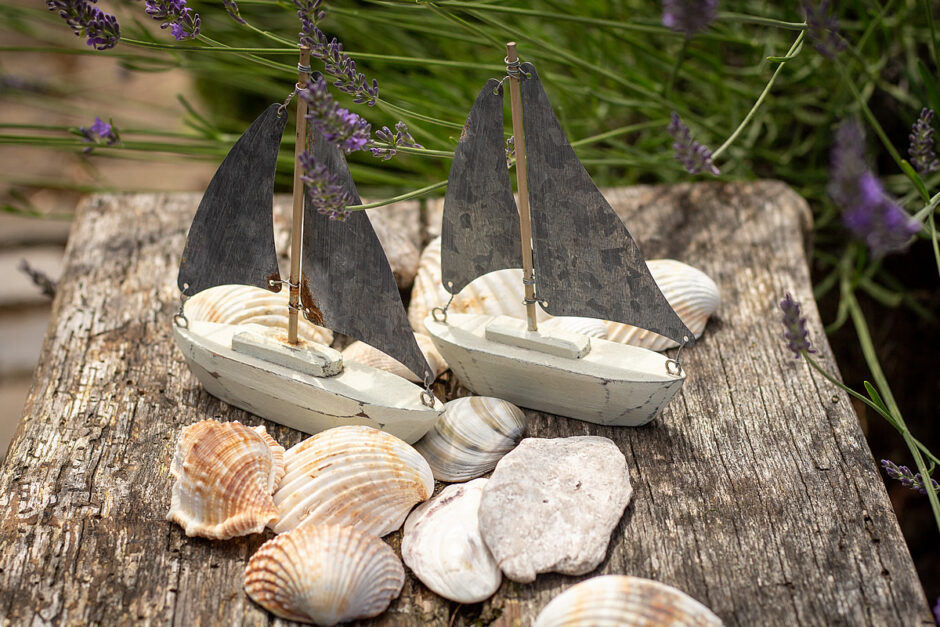 Ein sommerliches Deko-Arrangement aus zwei Segelbooten auf einem Holztisch und Muscheln davor.